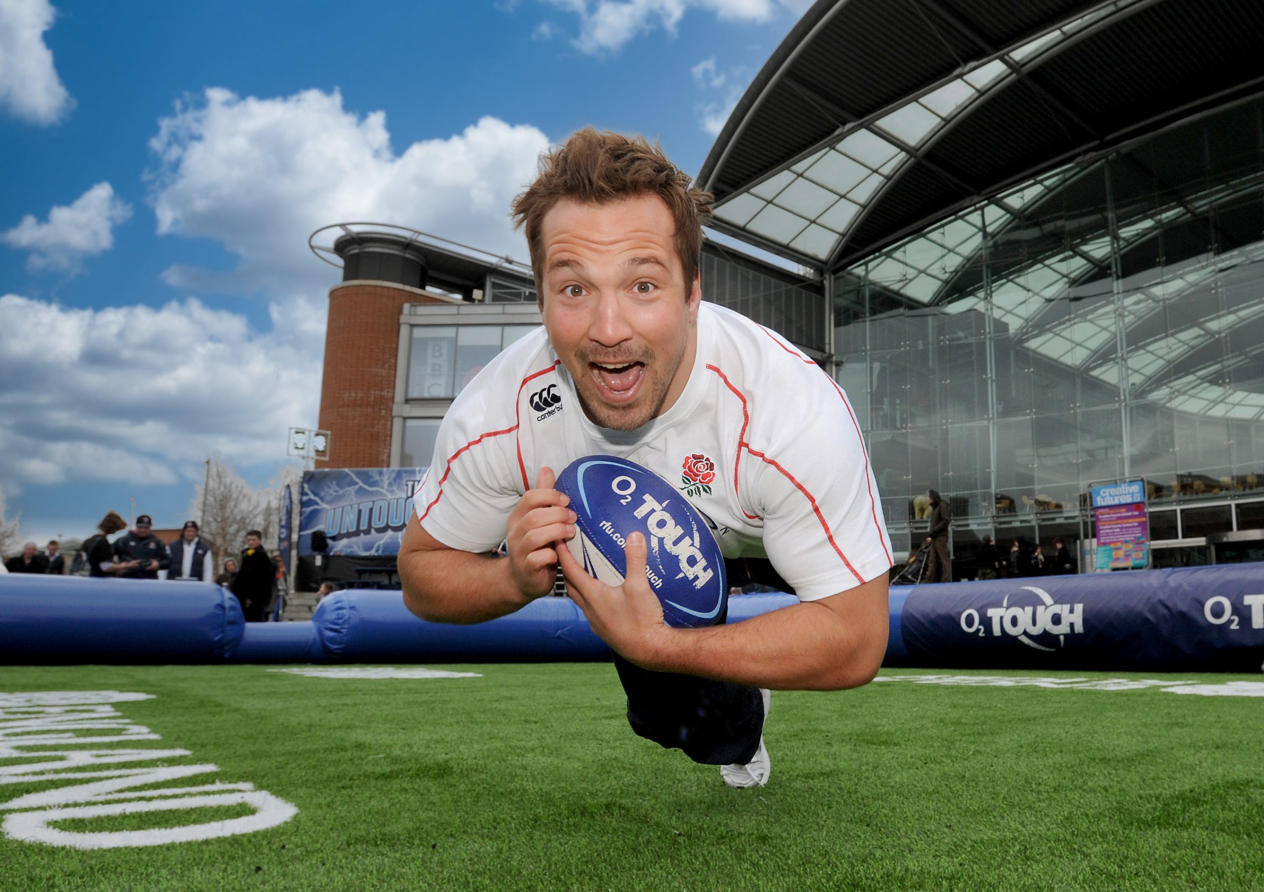 A man in a white rugby shirt dives forward holding a blue O2 Touch rugby ball on an outdoor artificial turf pitch, with a large modern building and a cloudy sky in the background.