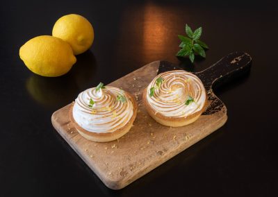 Two lemon meringue tarts with toasted tops sit on a wooden chopping board. Two whole lemons and a small sprig of mint are placed nearby against a black background.