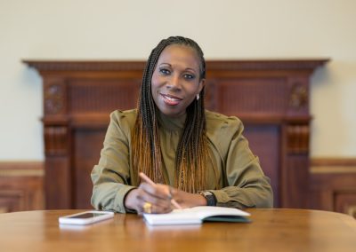 A woman with long plaits sits at a wooden table, writing in a notebook and smiling. A mobile phone is on the table beside her. A decorative wooden mantelpiece is in the background.