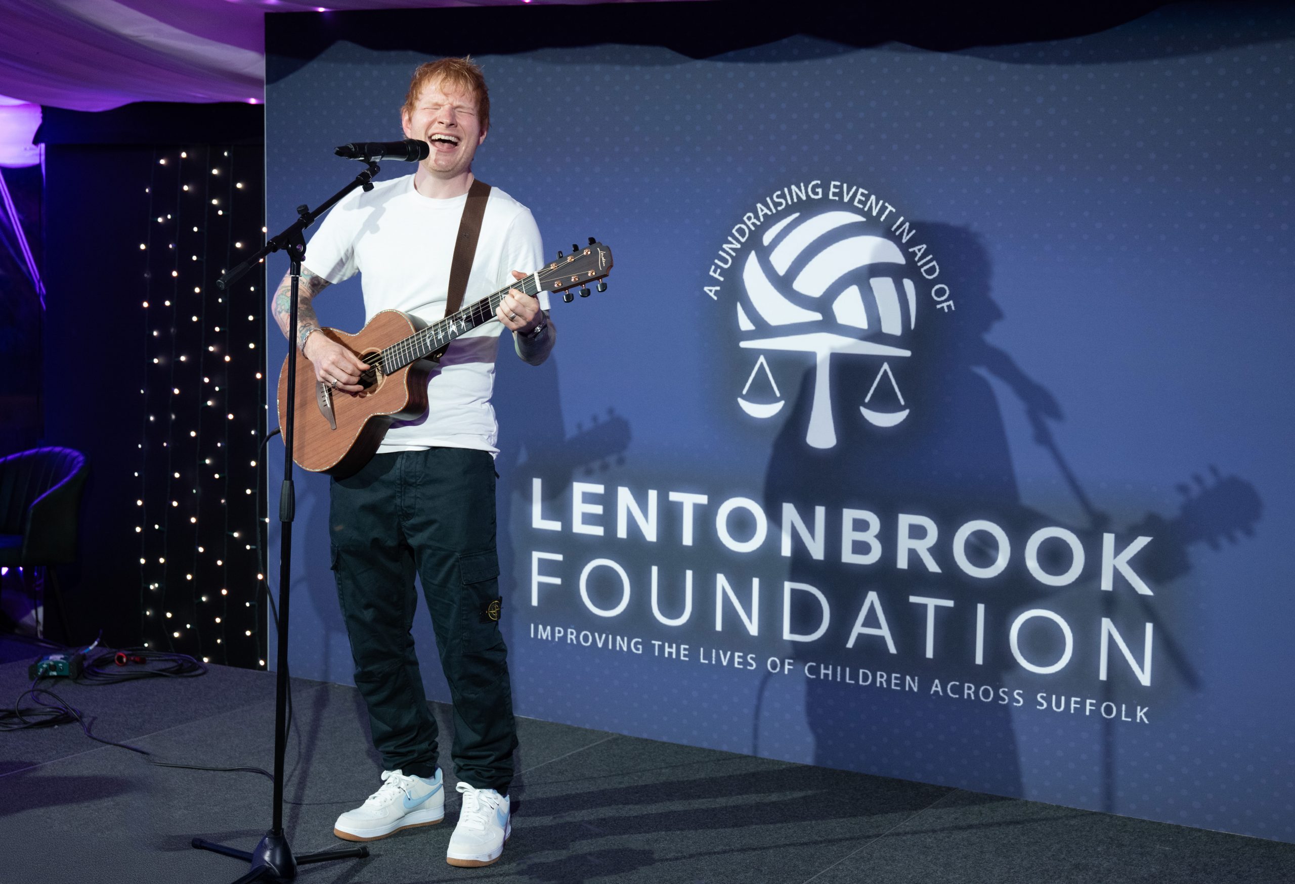 Ed Sheeran in a white T-shirt and jeans plays an acoustic guitar and sings on stage at a Lentonbrook Foundation fundraising event, with a blue backdrop displaying the foundation’s logo and slogan.